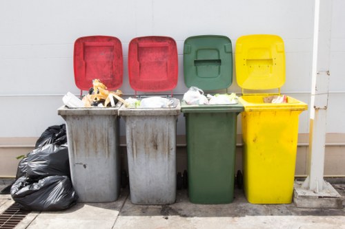 The image displays four large plastic wheeled bins placed in a row against a white wall. From left to right, the bins are in red, red, green, and yellow colours. The grey and red bins contain mixed waste, including white plastic bags, cardboard, and other rubbish visible at the top. The green bin appears to be empty or contains minimal waste, while the yellow bin has some small items and debris on its lid. To the left, there are several tightly tied black plastic rubbish bags stacked on the ground. The scene is set outdoors on a paved surface in a residential or commercial area, possibly in Clapham or nearby, with natural daylight illuminating the scene. The presence of a drainage grate on the ground indicates an environment suited for waste collection. The image is related to rubbish removal services offered by Office Clearance Clapham, illustrating bins prepared for rubbish collection or disposal.