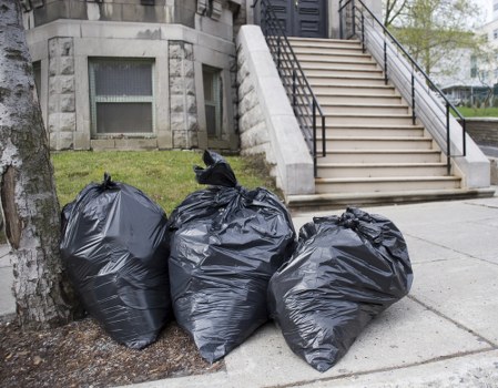 Three large black rubbish bags made of durable plastic are placed on the pavement near a tree, with a building and a set of external stairs in the background. The bags appear filled with assorted waste materials, and they are stacked closely together on a grey concrete sidewalk, adjacent to a grassy area and a stone base of the building. The building features a stone facade with small windows, while the metal staircase with white steps and black railings leads up to an entrance. The scene is well-lit by natural daylight, with some greenery visible in the background beyond the stairs, indicating an urban environment consistent with local rubbish removal services in Clapham. The image depicts a typical scene for waste collection and clearance activities undertaken by companies such as Office Clearance Clapham.