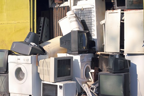 A pile of discarded electronic waste and household appliances arranged outdoors, including several old computer monitors with dark screens and beige plastic casings, washing machines with white enamel finishes, microwave ovens, and a small refrigerator or freezer. The appliances are stacked and leaning against each other, with some showing signs of wear such as dirt, scratches, and minor dents. The background features a partially visible yellow wall or fencing and a section of an industrial or storage area, with additional metal objects and debris nearby. The scene suggests a collection point for rubbish removal services in Clapham, where bulky waste items are temporarily stored before collection or disposal. Lighting appears natural, with shadows indicating daylight.

This image exemplifies typical rubbish removal activities, especially for bulky household waste, coordinated by companies like Office Clearance Clapham in the Clapham area, serving local needs for efficient clearance and recycling of electronic and appliance refuse.
