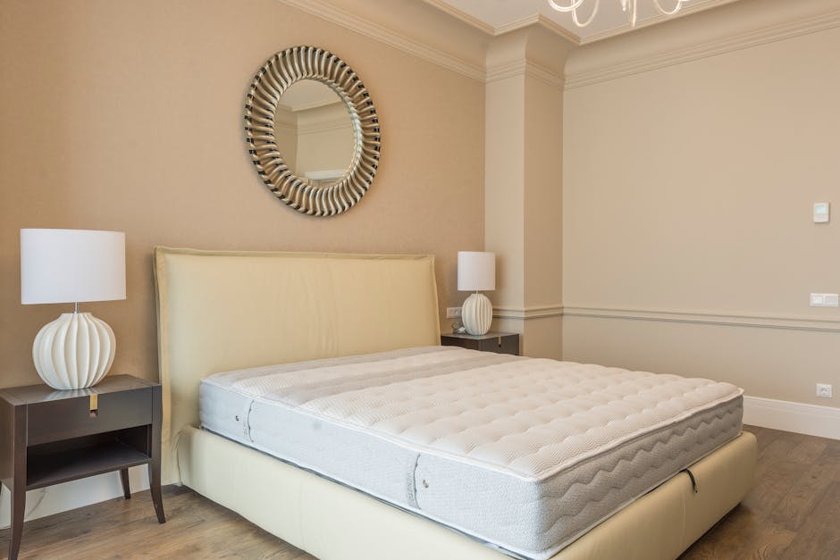 A minimalistic bedroom with white walls and ceiling, featuring a large white upholstered bed with vertical tufted detailing on the headboard and a plain mattress resting on a simple frame. The room ha