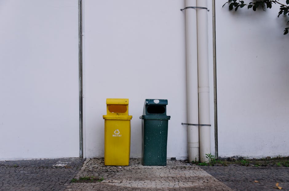 In front of a plain white exterior wall, two upright recycling bins are positioned side by side on a paved surface. The bin on the left is bright yellow with a rectangular opening at the top, labeled